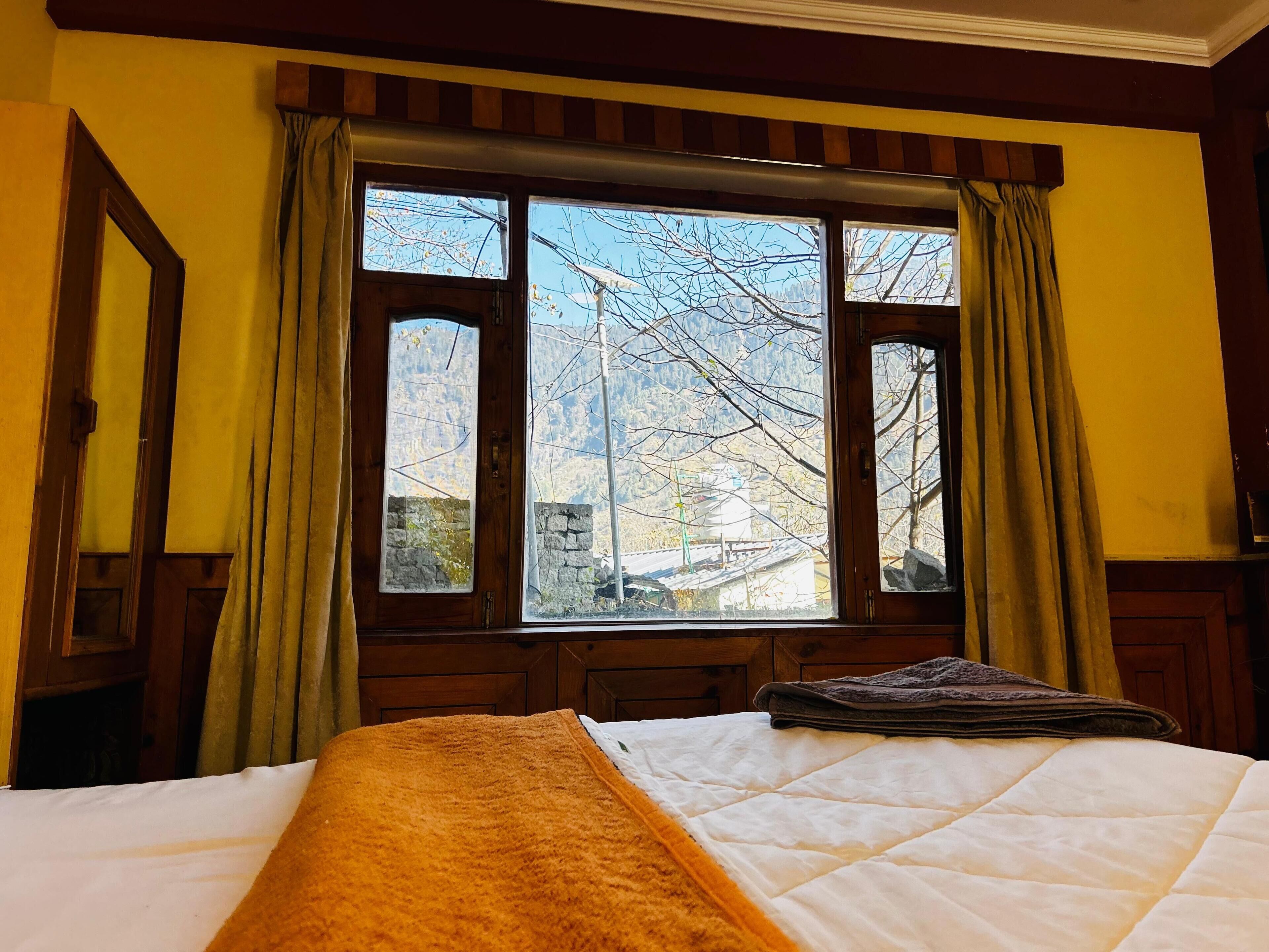 A room with a bed, a window with curtains, and a view of trees and buildings.