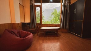 A room with a wooden floor, a red bean bag chair, a small table, and a view of green trees and mountains through a window with curtains. - Hostelam The Mountain Story (Manali)