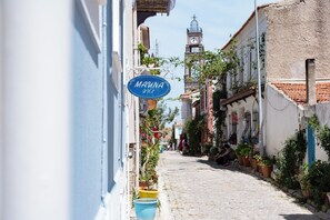 Une ruelle étroite pavée, bordée d’immeubles, un clocher visible au loin et une enseigne bleue indiquant « MAUNA HOTEL ».