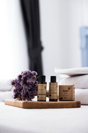 Trois bouteilles de savon sont posées sur un plateau en bois, avec une fleur violette, un rideau noir et une serviette blanche en arrière-plan.