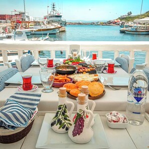 A table set with a variety of foods and drinks, overlooking a marina with boats. - Mauna Otel (Bozcaada)