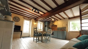 Een ruime woonkamer met een houten plafond, een eettafel met stoelen, een keuken met fornuis en kastjes, en een bank met turquoise kussens.