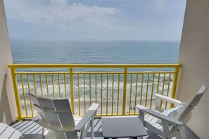 Een balkon met een gele reling, witte stoelen en een klein tafeltje.