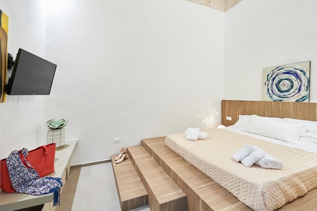 A hotel room with a bed, a TV mounted on the wall, a small table with a plant, and a red handbag on the floor.