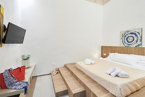 Ein Hotelzimmer mit einem Bett, einem an der Wand befestigten Fernseher, einem kleinen Tisch mit einer Pflanze und einer roten Handtasche auf dem Boden.