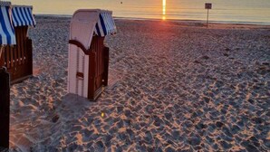 Ein Strand mit zwei Holzkufen, die mit gestreiften Tüchern abgedeckt sind, freier Blick auf den Sonnenuntergang über dem Meer und ein Schild in der Ferne.