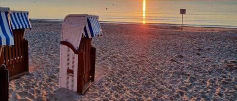A beach with two wooden chairs covered with striped cloths, a clear view of the sunset over the ocean, and a sign in the distance.
