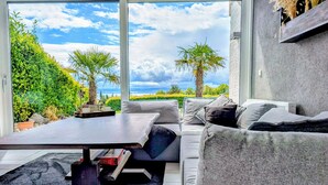 A modern living room with a large window, a grey sofa, a small table, and a view of the outdoors. - Vacation apartment with a view of Lake Constance under palm trees (Daisendorf)