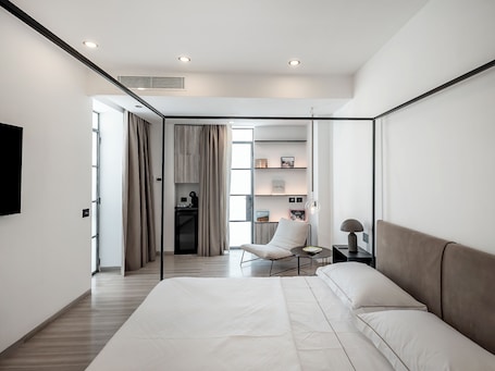 A modern bedroom with a large bed, a desk, and a chair. There is a bookshelf with books and decorative items.