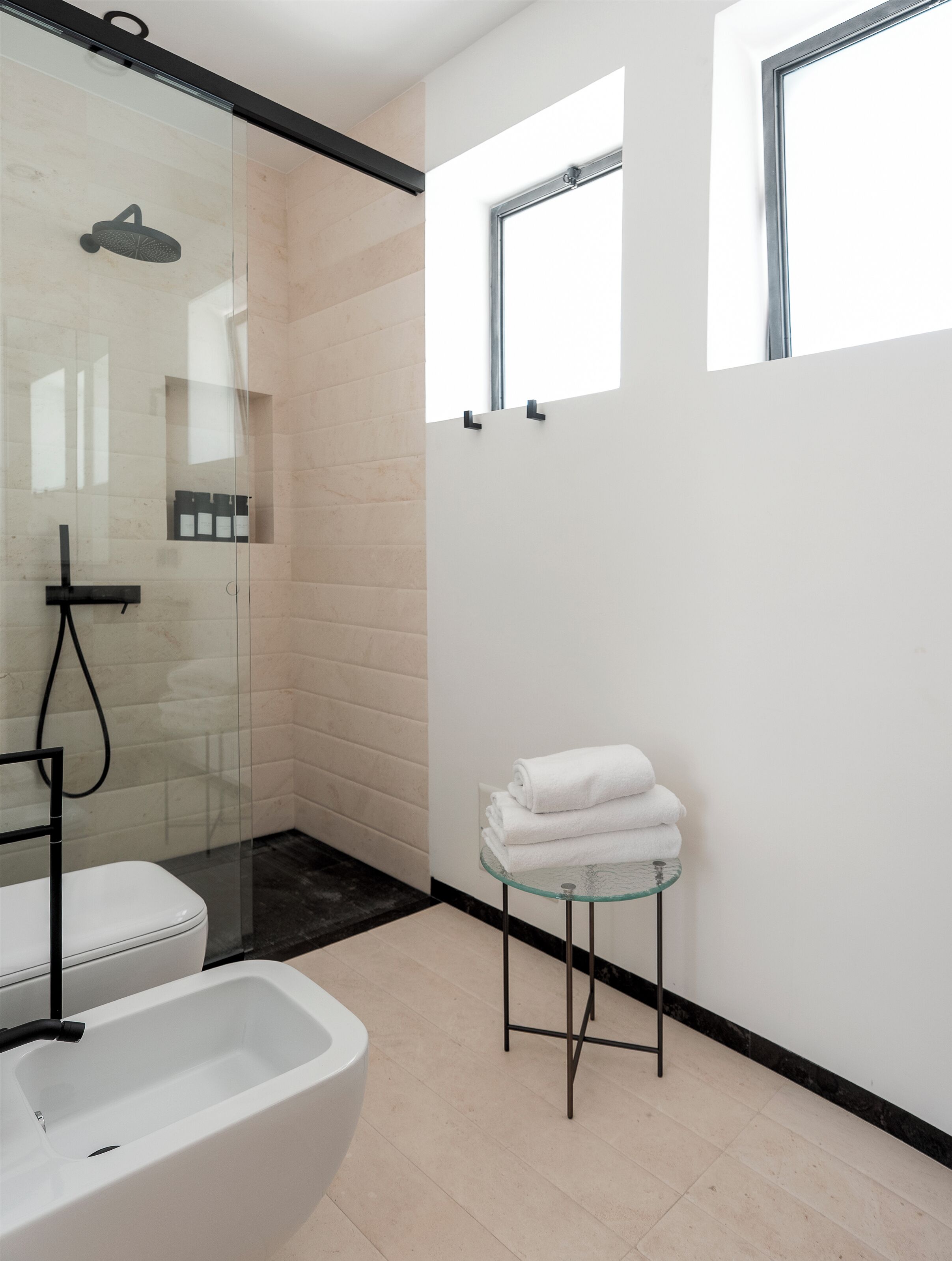 A modern bathroom with a glass shower enclosure, a white toilet, and a glass-topped vanity with towels.