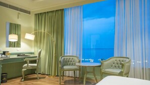 A hotel room with a large window, a desk with a chair, a round table with two chairs, and a bed.