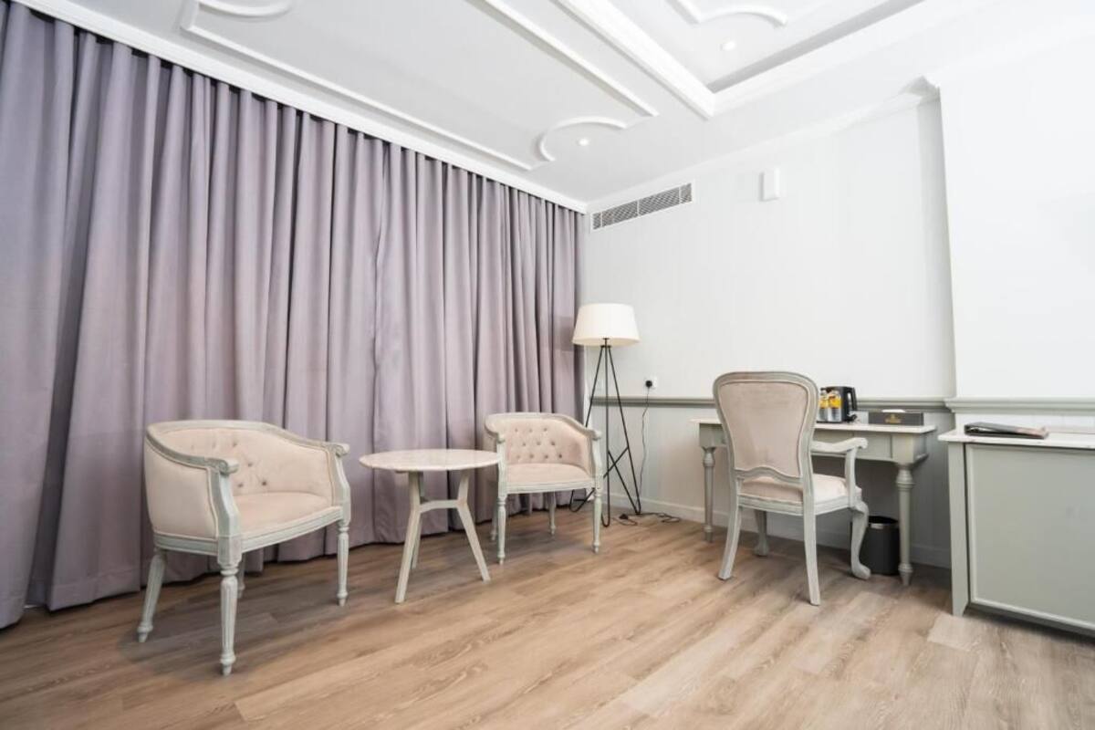 Quarto de hotel com piso de madeira, escrivaninha, duas cadeiras, luminária e uma janela grande com cortinas.