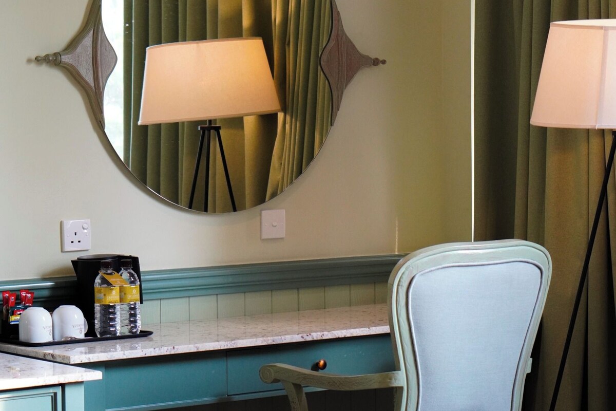 A room with a teal-colored desk, a round mirror with a decorative frame, a chair, a floor lamp, and a trash can.