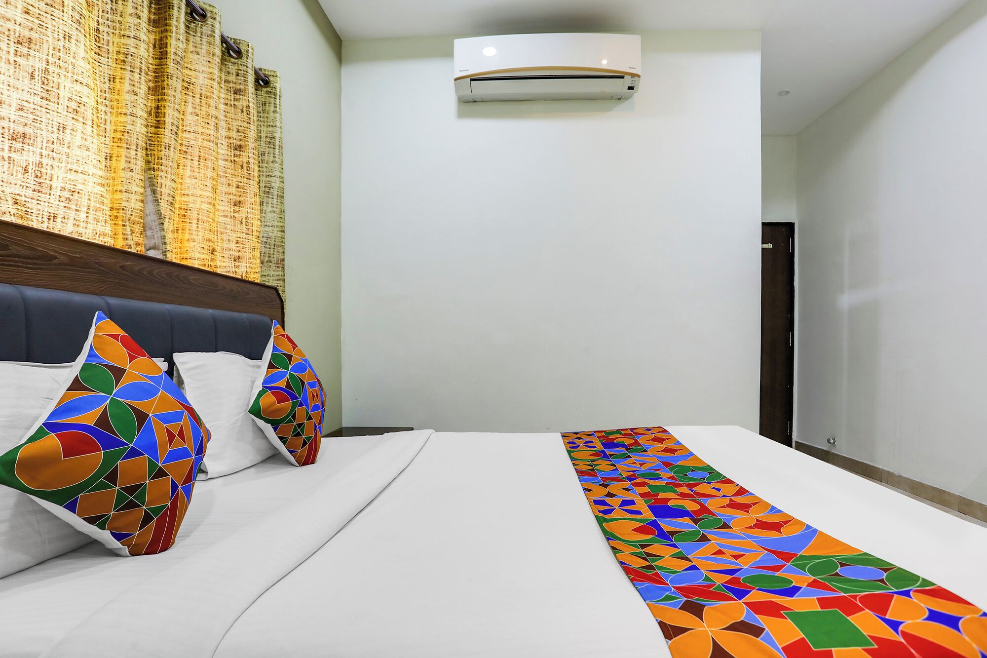 Quarto de hotel com cama, almofadas coloridas, cabeceira branca, ar condicionado fixado na parede e parede lisa.