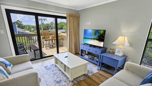 A living room with a TV, sofa, coffee table, and a view of a balcony with outdoor furniture. - 2BR Ocean Bridge - Shore Drive,Pool,Steps to Beach (Myrtle Beach)