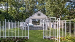 A small house with a porch, surrounded by a white fence and lush greenery.