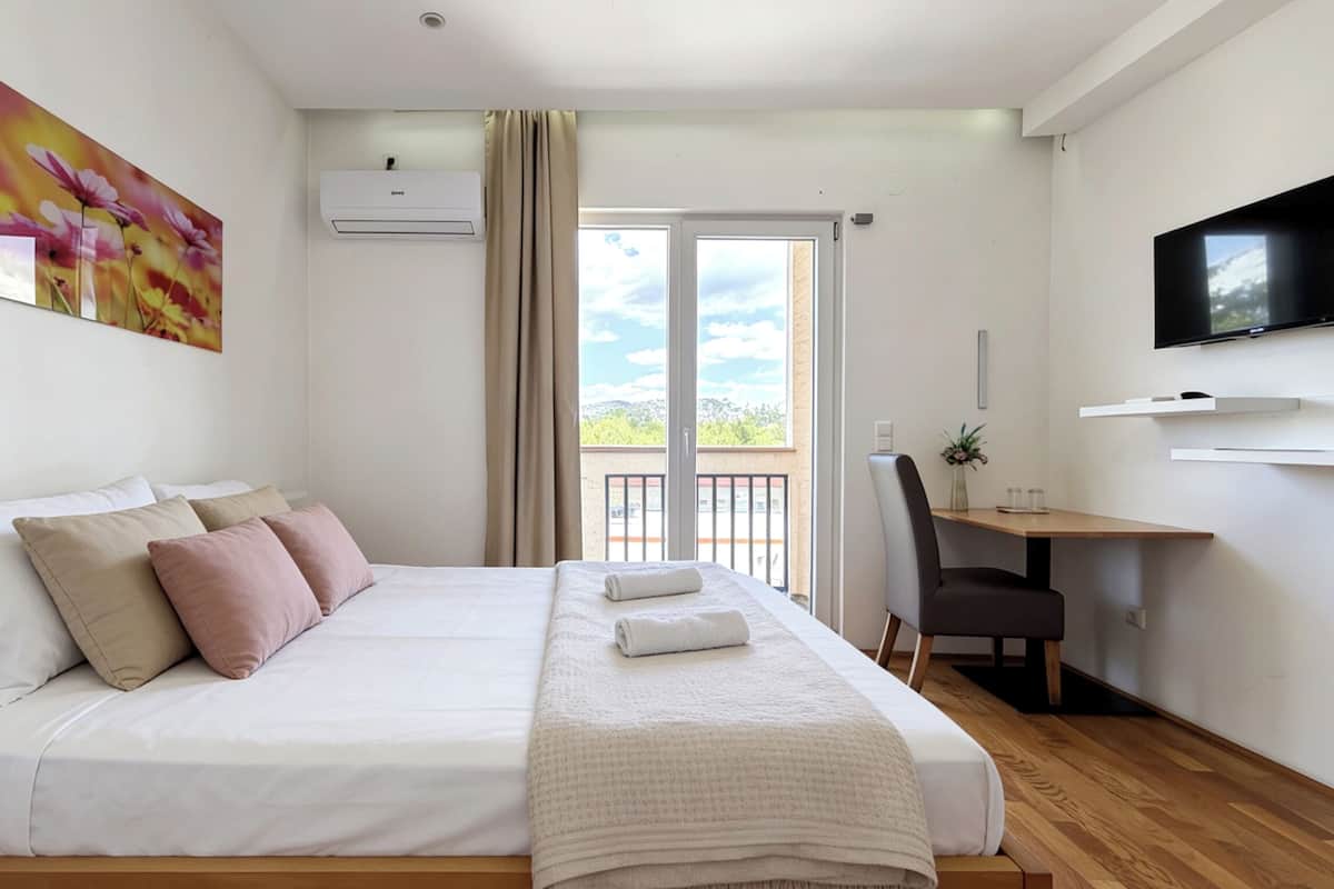Habitación de hotel con cama, escritorio, silla, televisor y balcón con vistas.