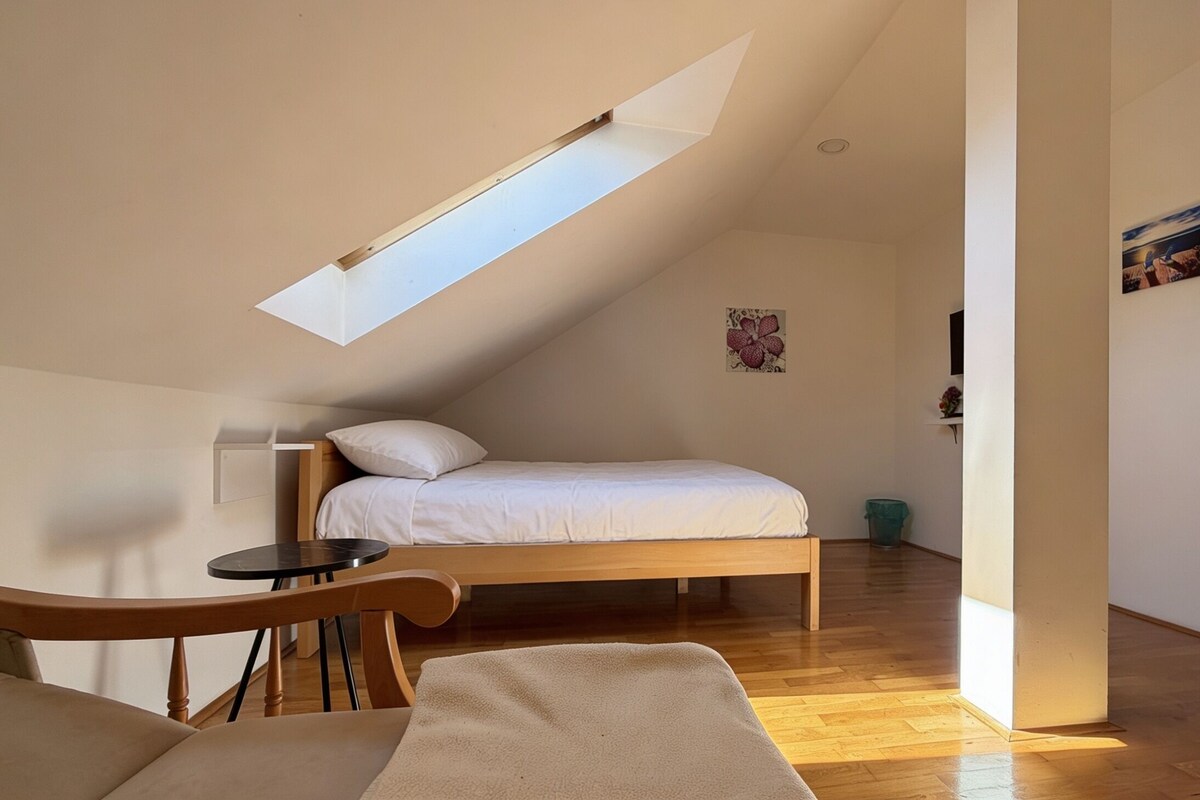 Un dormitorio con techo inclinado, una cama, una mesita pequeña y una silla.