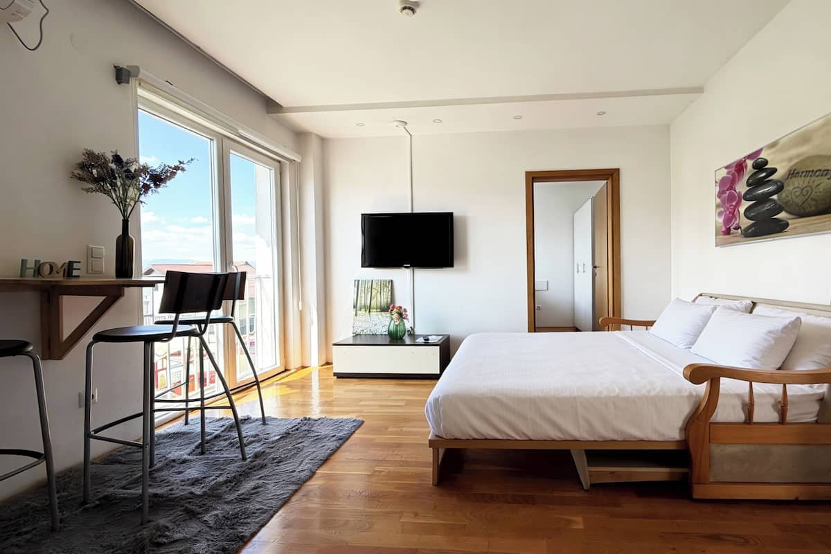 Un dormitorio moderno con una cama de madera, un escritorio con una silla, un televisor y un ventanal grande.