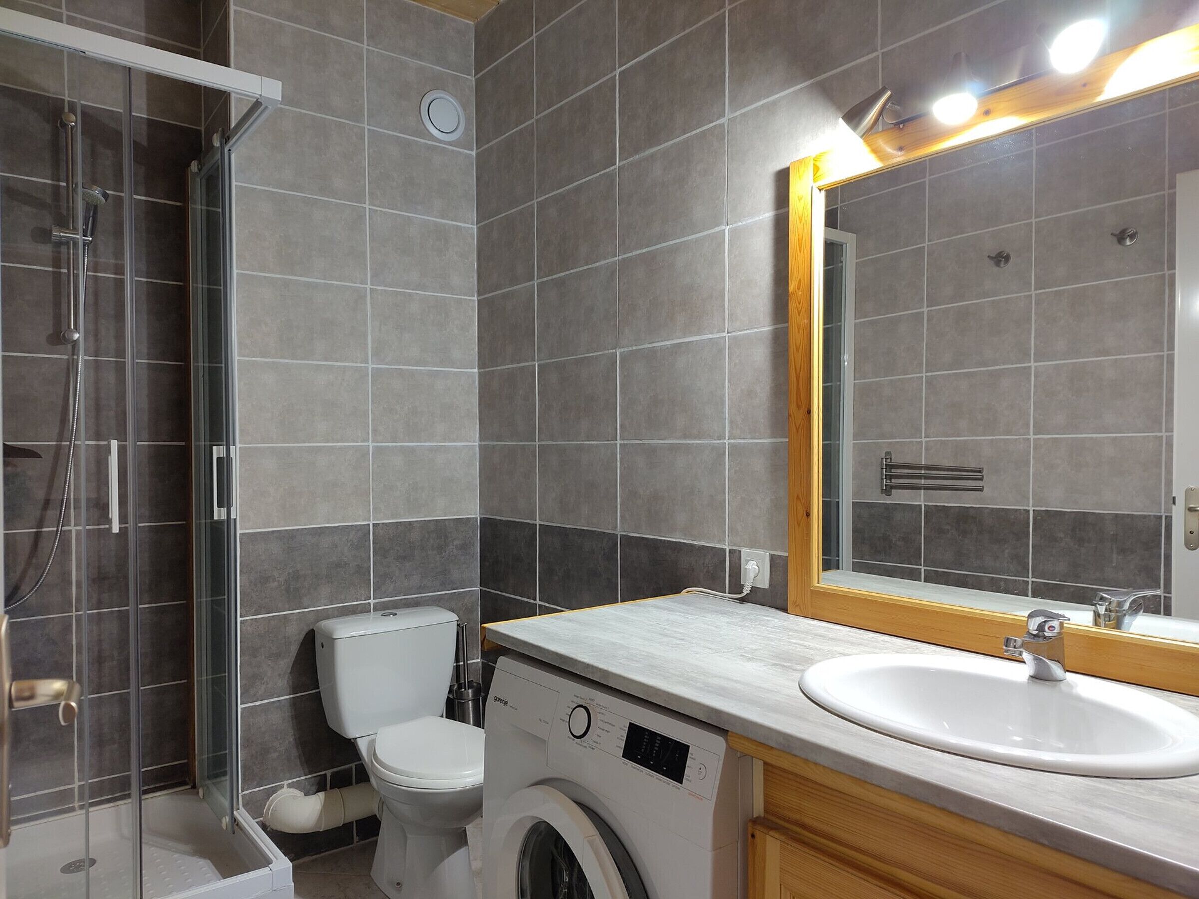 A modern bathroom with a glass shower enclosure, a toilet, a washing machine, and a large mirror with a wooden frame.
