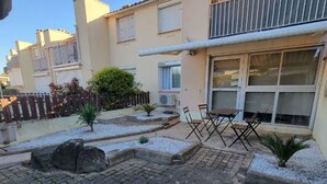 Un edificio residenziale con un patio arredato da tavolo e sedie, un piccolo giardino con piante e rocce, e un recinto in legno.