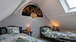 Une chambre avec deux lits, chacun recouvert d’une couvre-lit orné de motifs de feuilles tropicales, dotée d’un ventilateur fixé au mur et d’une lucarne.