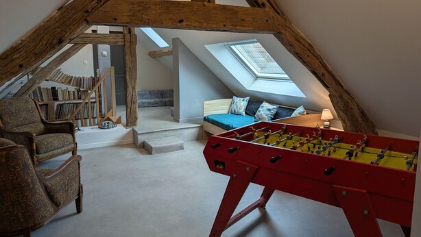 Une chambre à coucher aménagée dans un espace de type « loft », avec un plafond haut, des poutres apparentes, une table de baby-foot, un canapé et une lucarne.