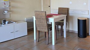Un ambiente compatto con angolo ristoro, un mobile bianco con cassetti, una sedia in vimini, un tavolino, un cestino nero e un quadro con pappagalli.