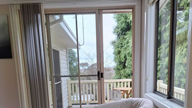Una habitación con sofá color beige, puerta corrediza de vidrio que da a un balcón y vistas a árboles y una valla.