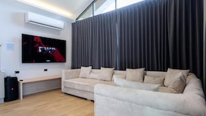 A modern living room with a beige sofa, a flat-screen TV mounted on the wall, and a large window with curtains. - Six elements pool villa khaokho (Khao Kho)