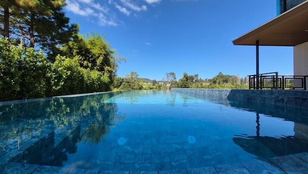 A clear blue swimming pool with a stone edge, surrounded by lush greenery and a modern building with a covered patio area. - Six elements pool villa khaokho (Khao Kho)