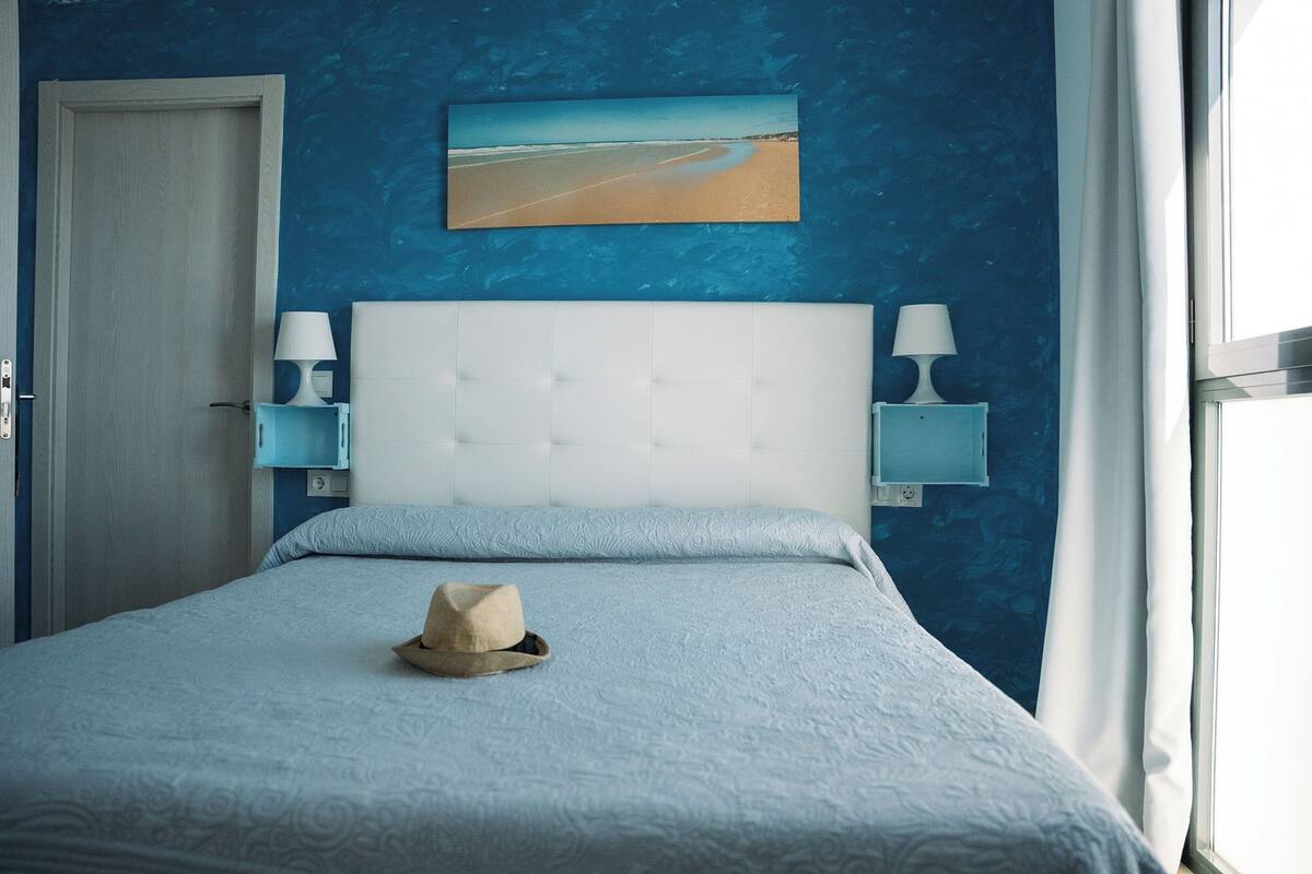 Um quarto com uma cama, mesa de cabeceira, luminária na parede, quadro e uma porta.