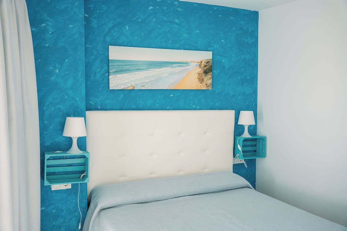 Quarto com parede azul, cabeceira branca e pintura com tema de praia.