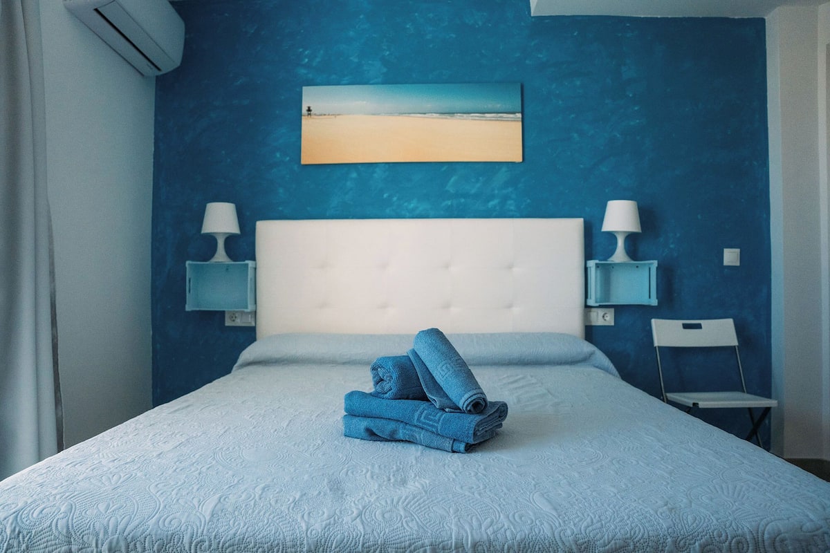 Um quarto com parede azul, cabeceira branca, duas luminárias de cabeceira, uma toalha dobrada na cama e um quadro de uma praia.