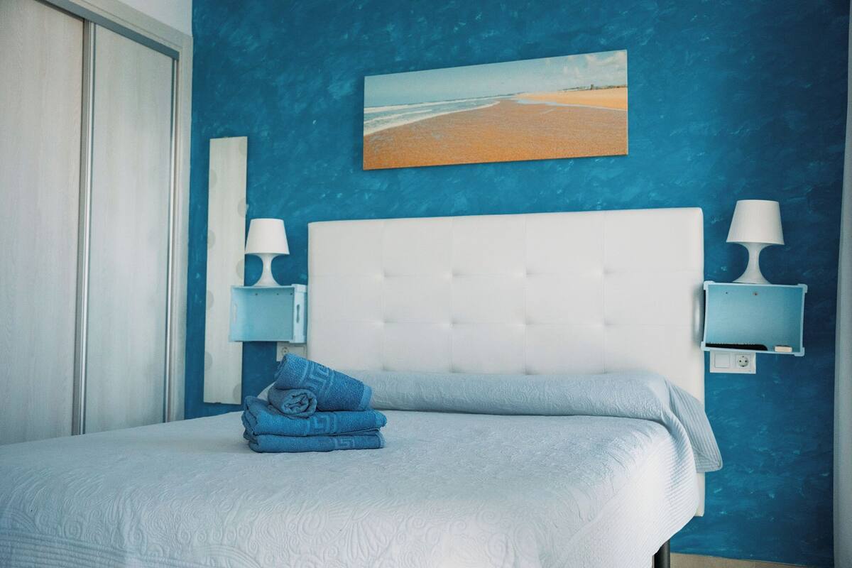 Quarto com cabeceira branca, duas mesas de cabeceira com luminárias, uma toalha azul dobrada e um quadro de uma praia.