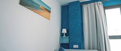 Un dormitorio con cama, un cuadro, una mesita de noche con lámpara y una ventana con cortinas.