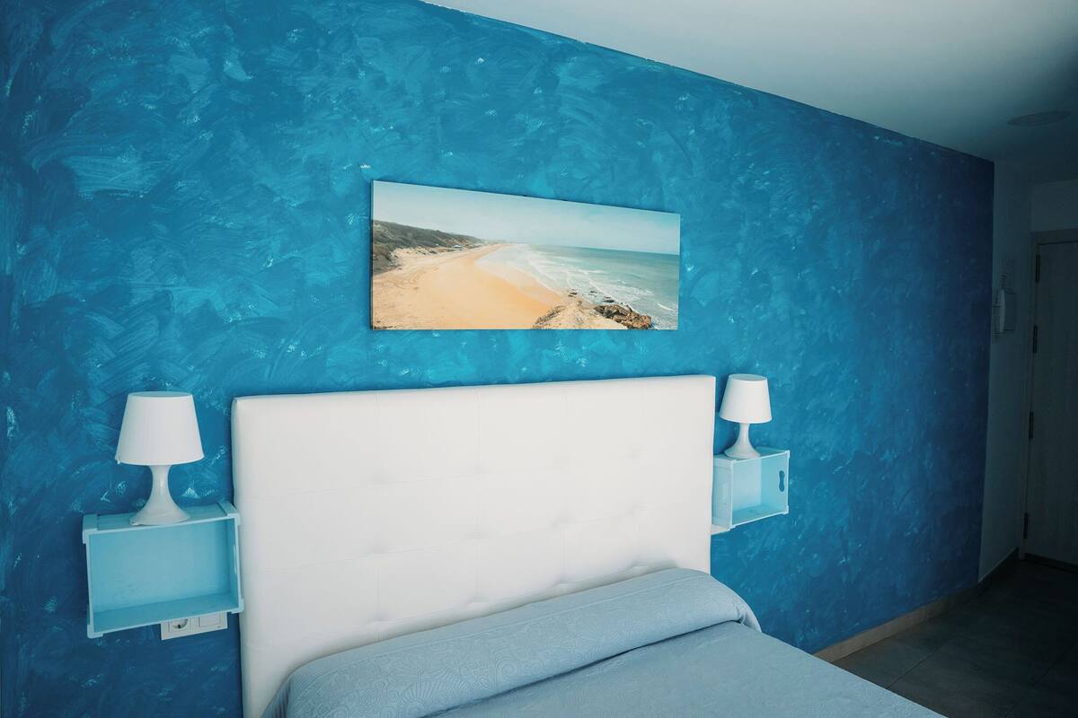 Um quarto com cabeceira branca, duas mesas de cabeceira com luminárias, um quadro com tema praiano na parede e uma porta ao fundo.