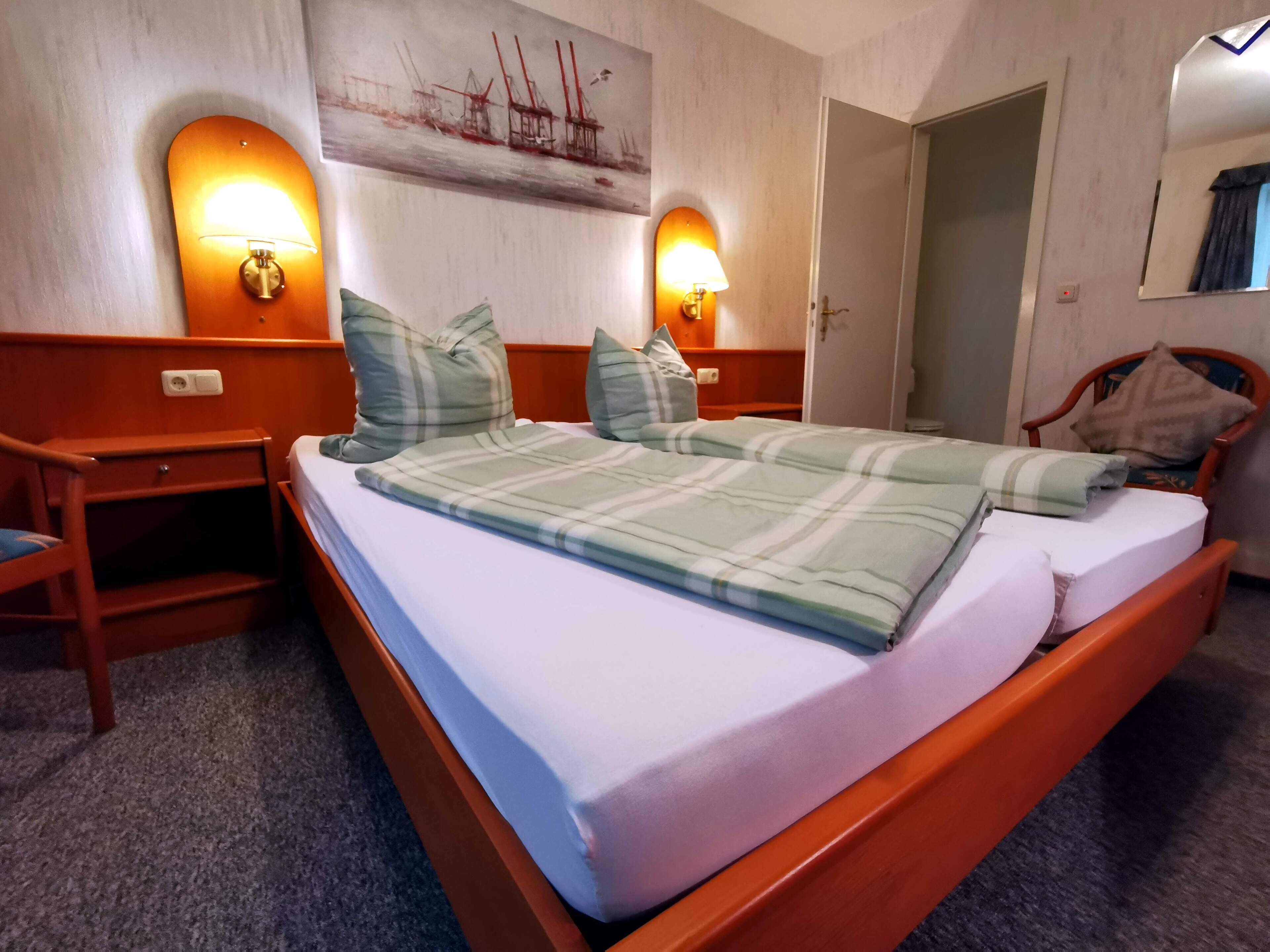 Una camera d'albergo con un letto di legno, due lampade sui comodini, un quadro appeso al muro e una poltrona con cuscino.