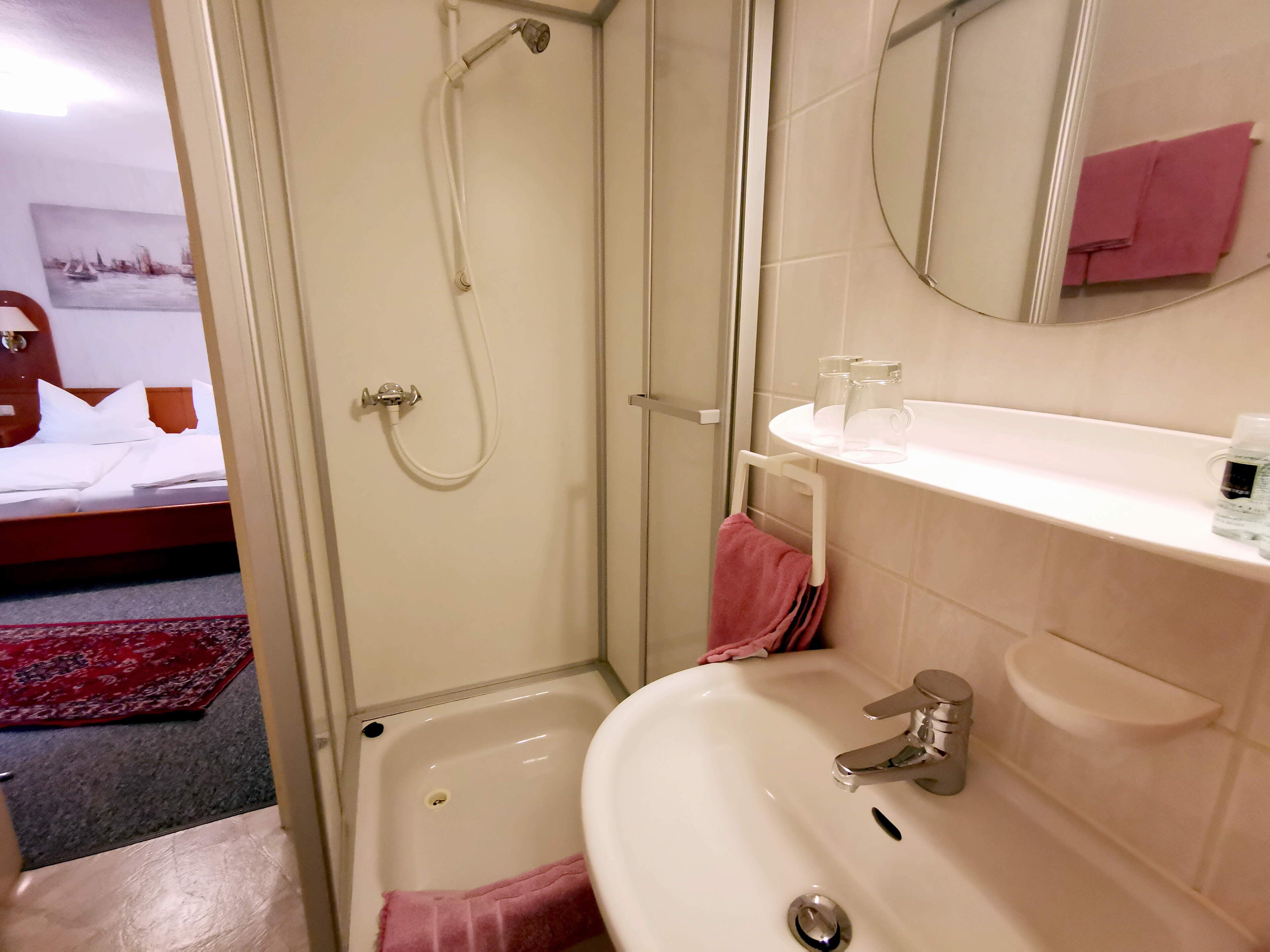 Un baño de hotel con ducha, lavabo y una cama visible al fondo.
