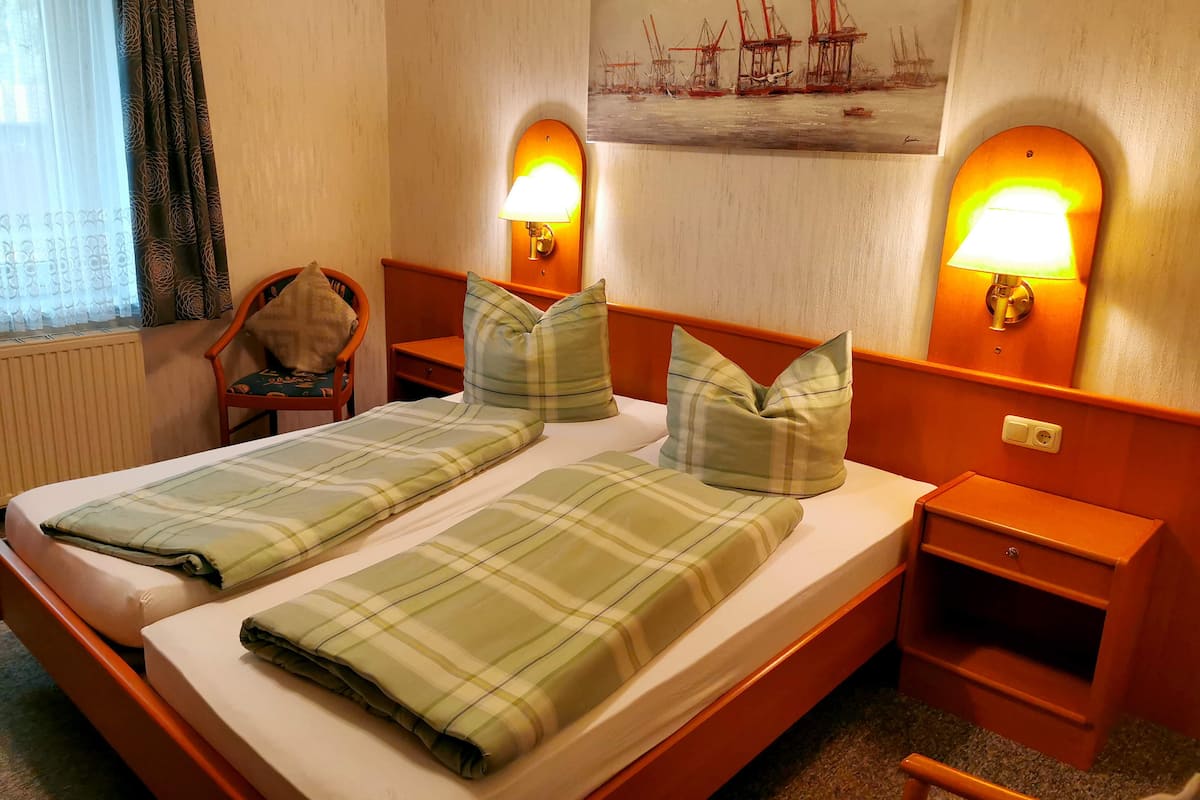 A double bed with plaid bedding, two bedside tables with lamps, a chair, and a painting of ships on the wall.