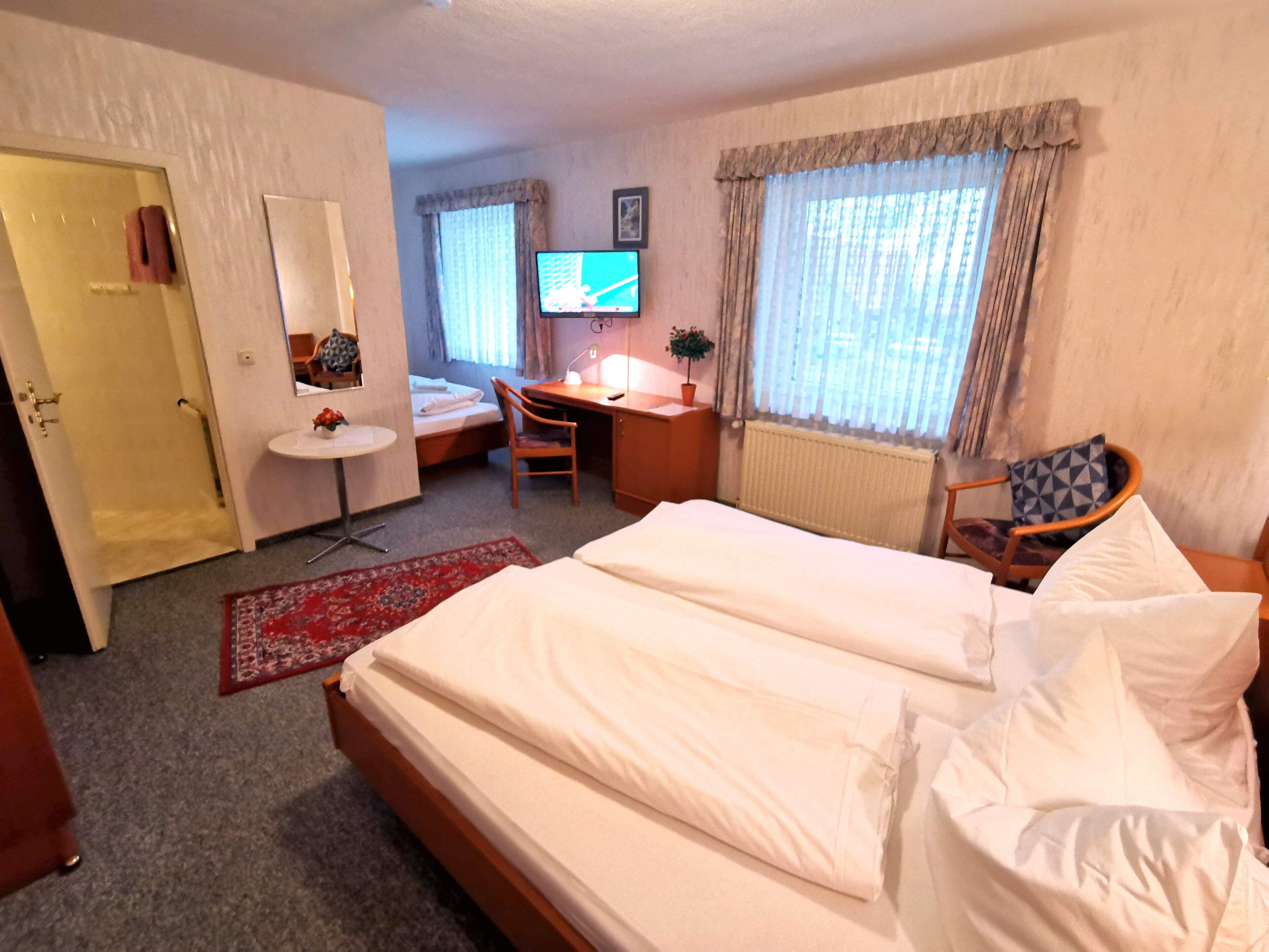 A hotel room with a bed, a desk, a chair, a small table, a mirror, a TV, and a window with curtains.