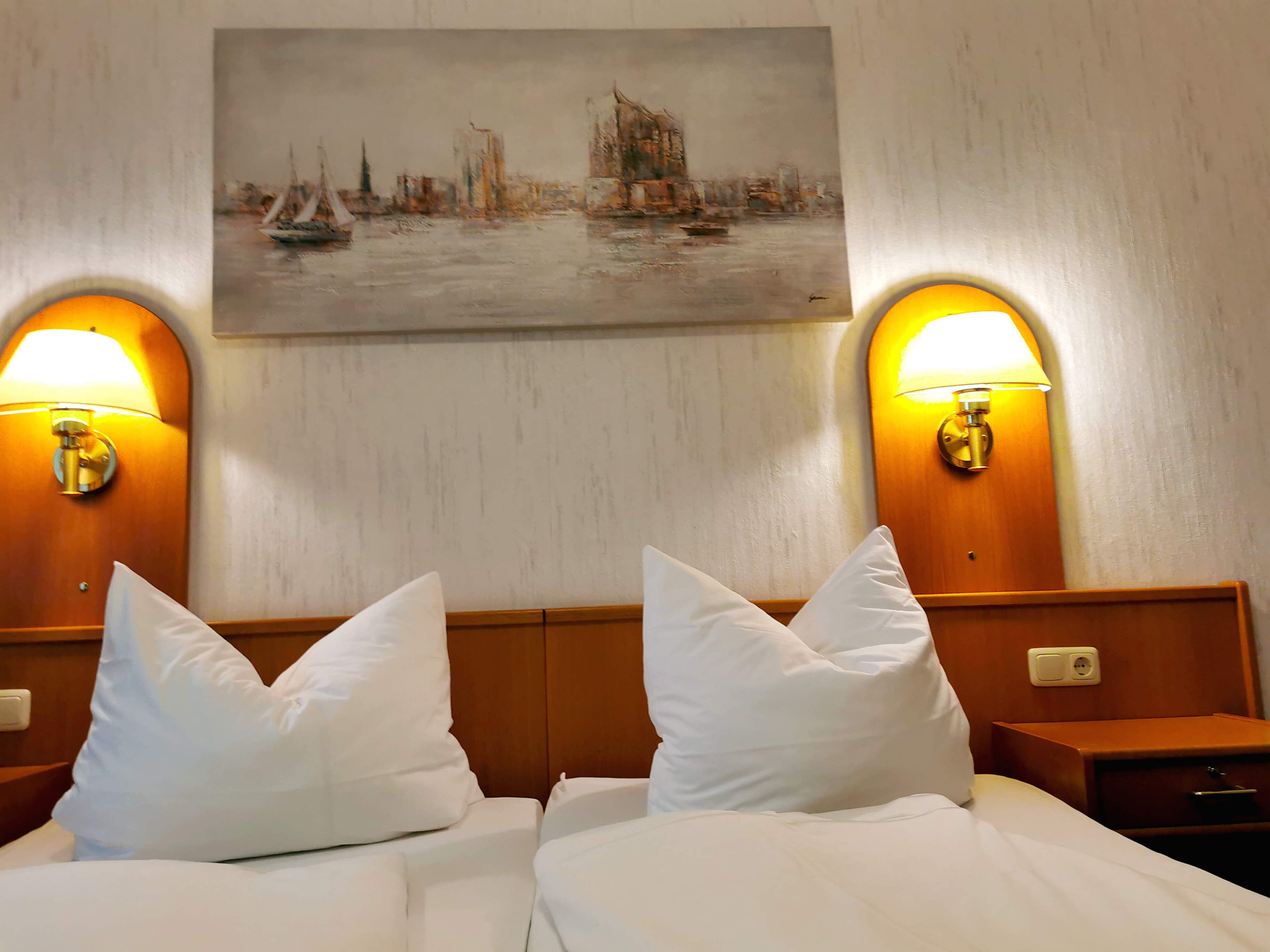 Quarto de hotel com duas camas, roupa de cama branca e duas luminárias fixadas na parede.