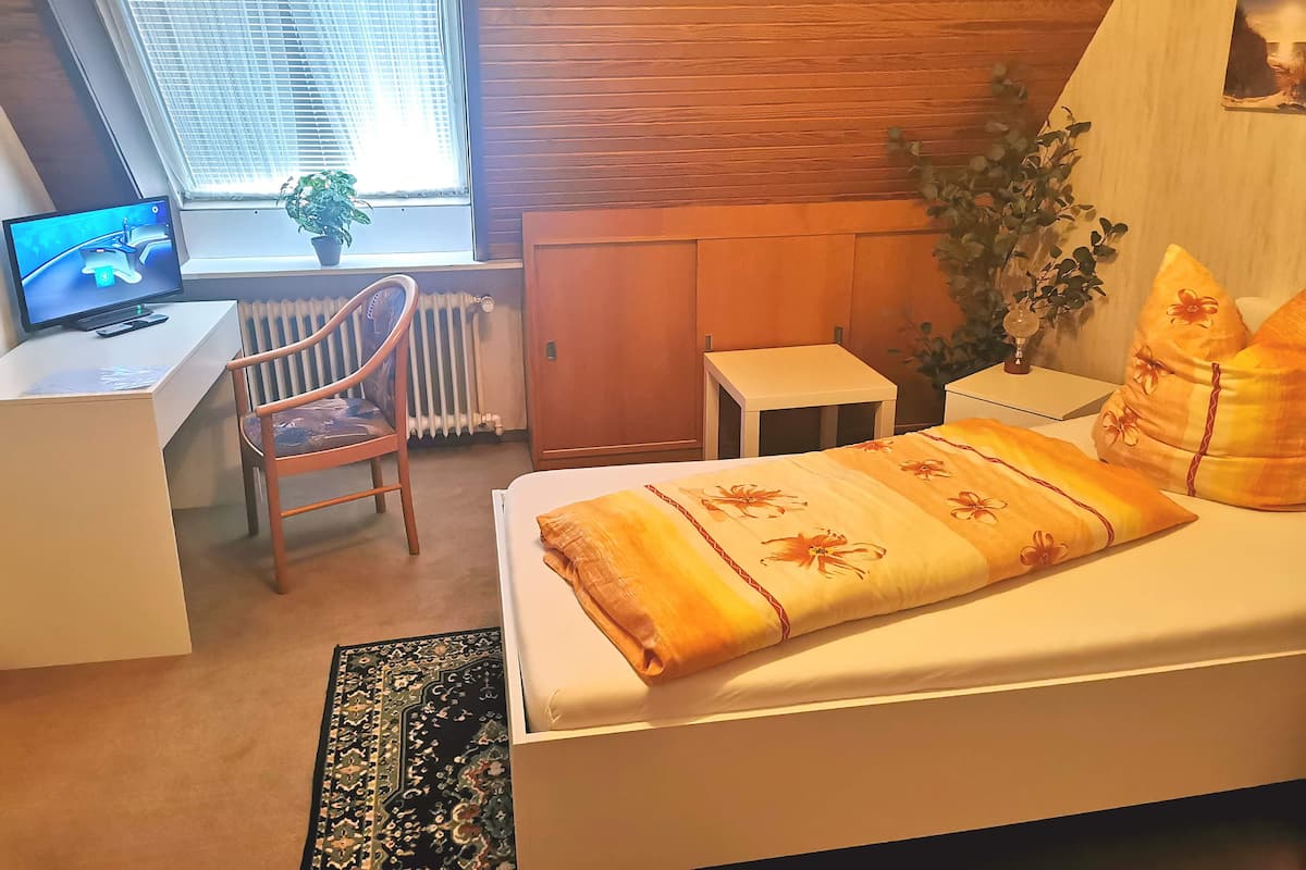 A bedroom with a bed, a desk, a chair, a radiator, a small table, and a window with blinds.