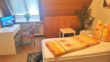 A bedroom with a bed, a desk, a chair, a radiator, a small table, and a window with blinds.