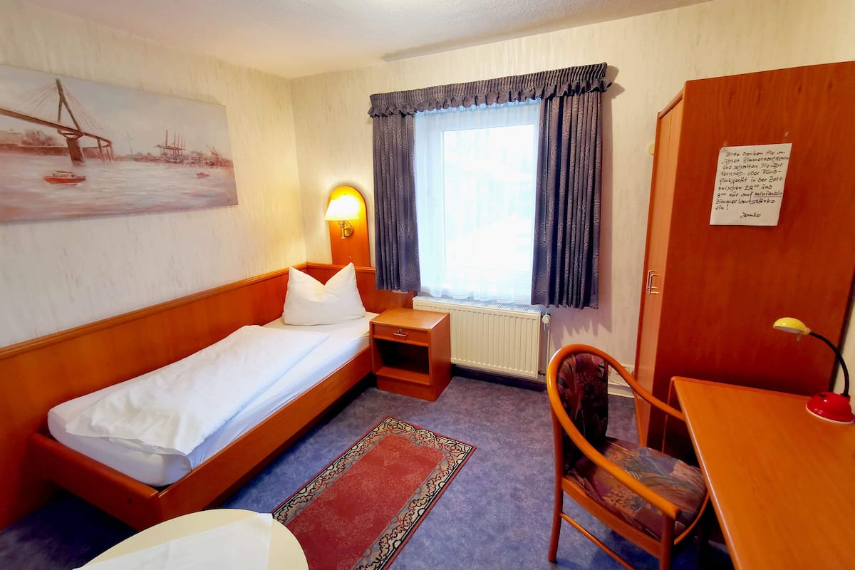 A single bed with a pillow, a bedside table, a chair, a desk, a lamp, a radiator, a window with curtains, a painting on the wall, and a note on the door.