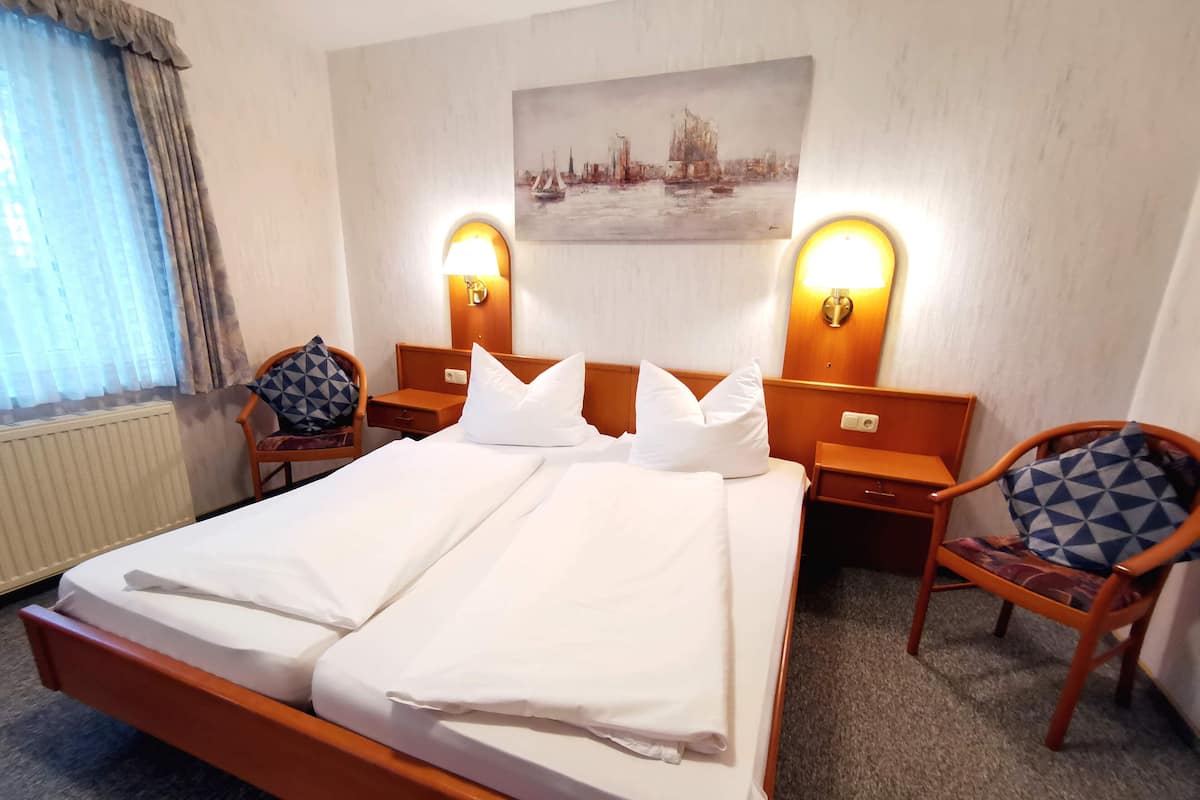 A hotel room with a double bed, two nightstands with lamps, a chair with a geometric-patterned cushion, and a painting on the wall.