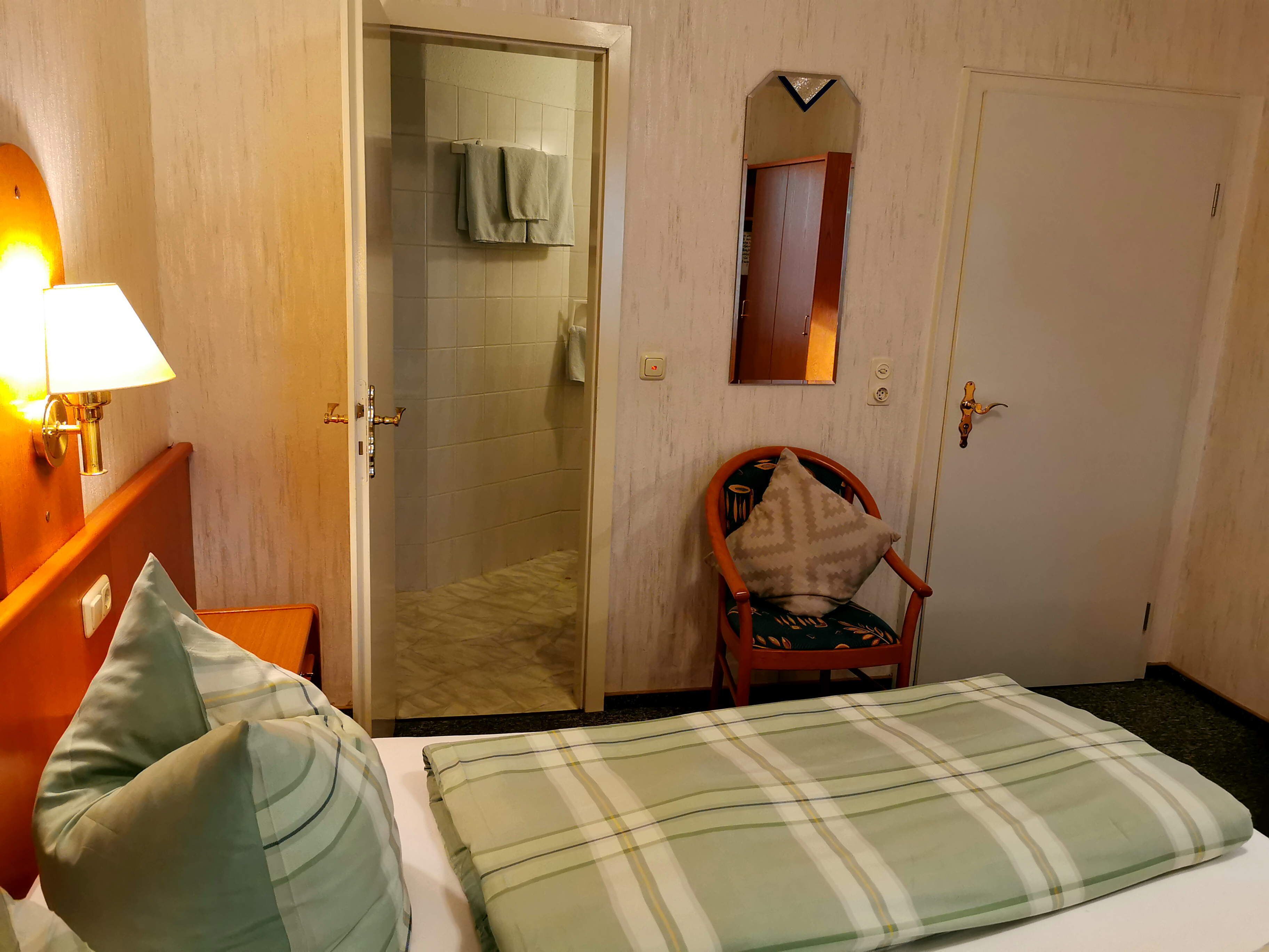 A hotel room with a bed, a chair, a bathroom with a towel rack, and a mirror on the wall.