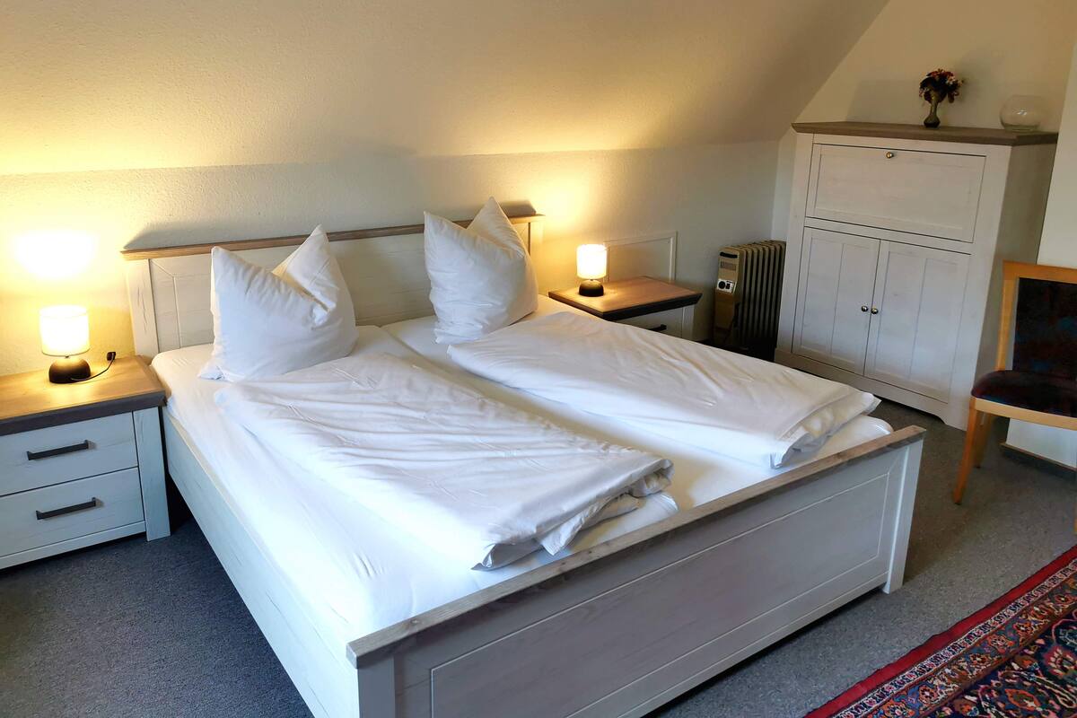 A neatly made bed with white linens, two bedside tables with lamps, a radiator, and a chair.