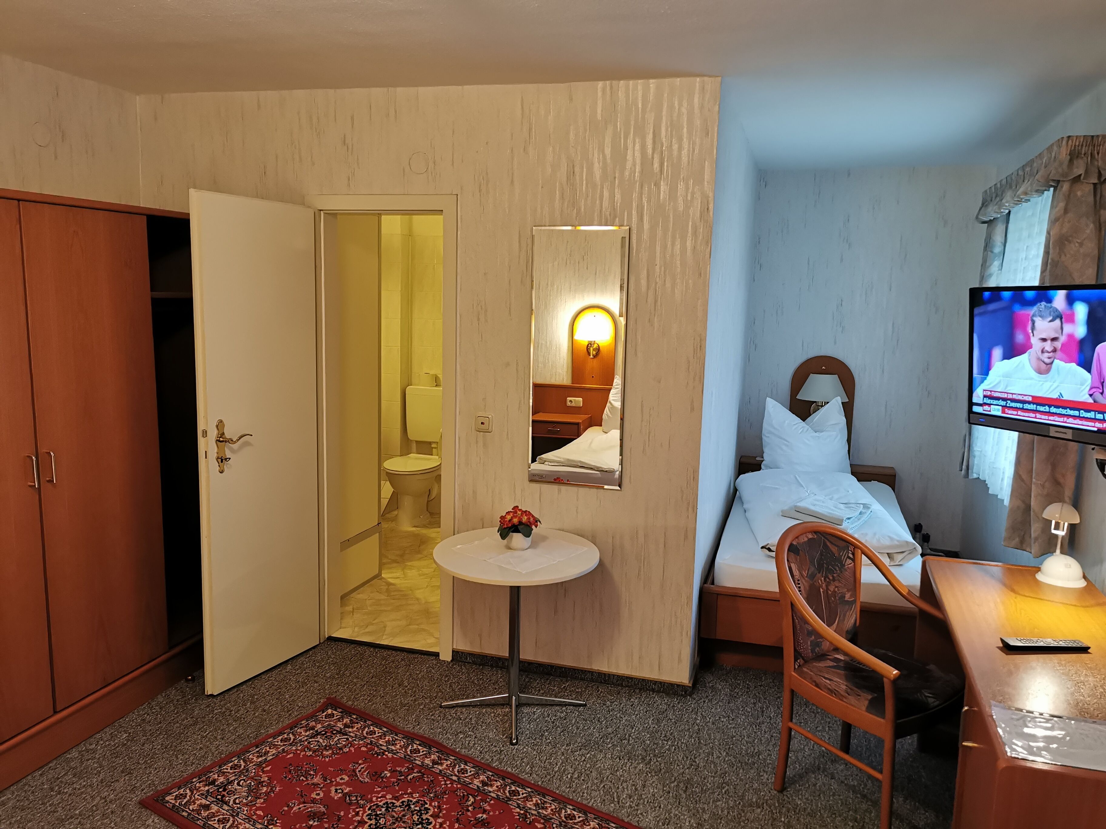 Habitación de hotel con cama, televisión, mesita, silla, escritorio, espejo y armario.
