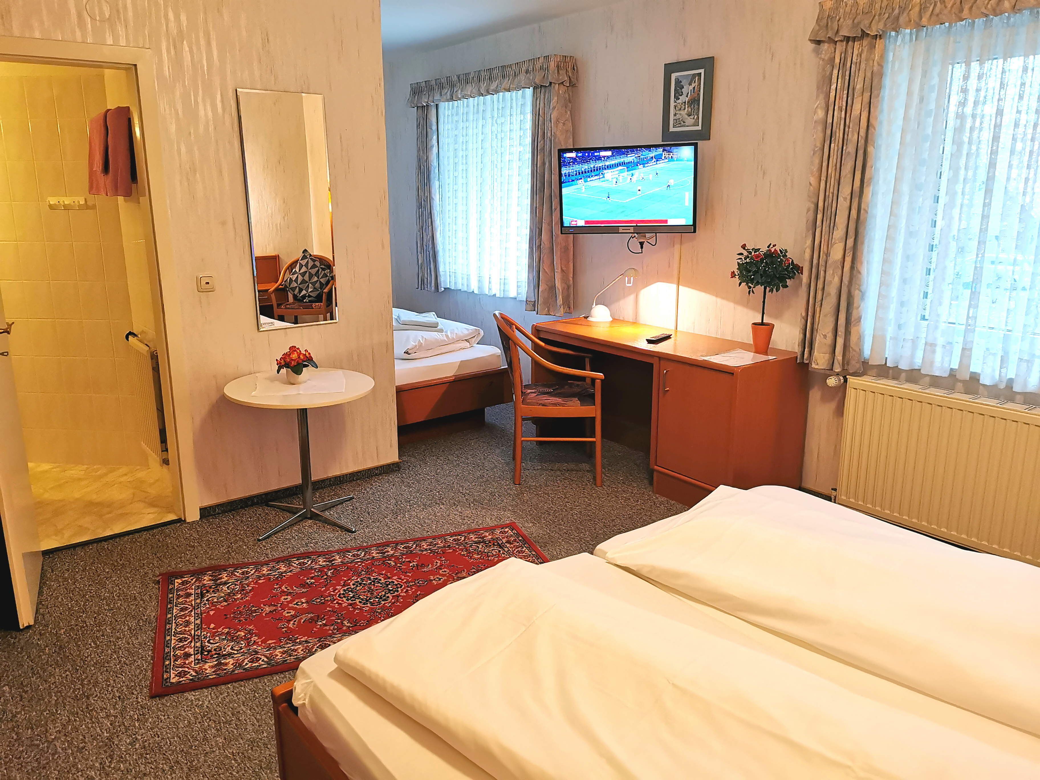 A hotel room with a bed, a desk, a chair, a small table with a vase, a TV, and a window with curtains.